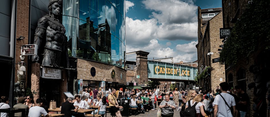 A busy outdoor scene in Fitzrovia, London, showing a crowded pedestrian street with numerous people sitting at tables outside cafes and restaurants. On the left side of the image, there is a large decorative statue of a figure in historical attire mounted on the building's brick wall. The buildings feature modern glass facades and traditional brickwork, with various signs and shop fronts visible, including a prominent hanging sign that reads 'Camden Lock.' Overhead, a dark green bridge structure spans the street, displaying a sign with the text 'Camden Lock.' The sky above is partly cloudy with patches of sunlight illuminating the scene. Some pedestrians are engaged in conversations, while others are walking or looking at their phones. This outdoor setting is amid a lively area suitable for social gatherings, and it captures the vibrant atmosphere characteristic of house removal and furniture transport logistics in Fitzrovia, as handled by [COMPANY_NAME].