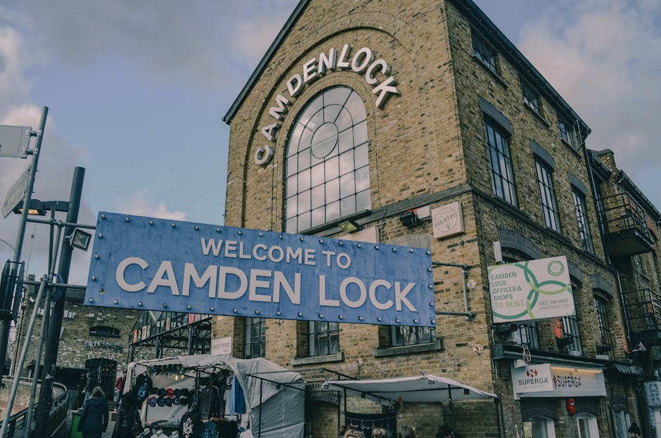 The exterior of a three-storey brick building in Fitzrovia, London, featuring a large arched window on the upper floor with the name 'CAMDEN LOCK' displayed prominently. In front of the building, there is a blue sign that reads 'WELCOME TO CAMDEN LOCK' attached to a black metal fence. The scene includes a small outdoor market stall covered with a white canopy, displaying various items such as clothing or accessories, with pedestrians nearby. Adjacent signage indicates rental services for Camden Lock Lock & Offices, and the street environment shows typical urban features like lampposts, other shops, and residential balconies, all under a partly cloudy sky. The setting appears to be during daytime with natural lighting. This scene relates to home relocation and furniture transport activities, as evidenced by the proximity of the market area and street-side setting. Man with Van Fitzrovia specializes in removals and moving services, including loading and transportation of household items, which may involve navigating such busy urban environments.
