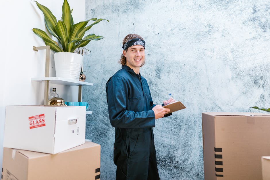 A person wearing a yellow t-shirt, blue jeans, and a wristwatch is standing indoors near several large cardboard boxes, some of which are open with flaps folded back. They are holding a clipboard in t