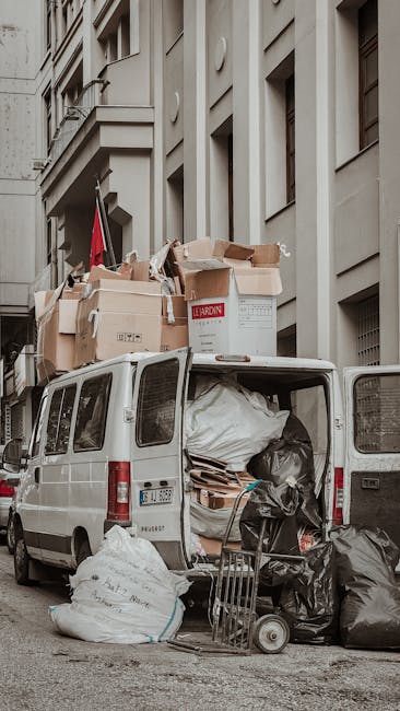 A white commercial van with its rear doors open, positioned on a city street in Fitzrovia, surrounded by large cardboard boxes, plastic-wrapped furniture, and black garbage bags loaded into the vehicle for a house relocation or bulky waste removal. The boxes are stacked on top of the van, some partially collapsed, with packaging materials visible. A hand truck or dolly is placed beside the open van, loaded with additional waste materials, ready for collection or transport. The background features a multi-storey building with beige, classical-style architecture and several windows. Natural daylight illuminates the scene, highlighting the process of loading waste or unwanted items as part of a professional removal or clearance service provided by Man with Van Fitzrovia, supporting efficient moving logistics and packing and moving operations.