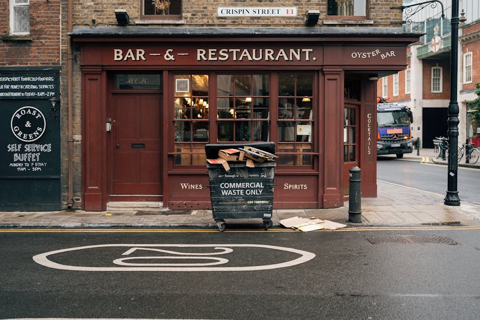 The image shows a street scene in Fitzrovia featuring a brick-fronted building that houses a bar and restaurant, with signs indicating offerings of wines, spirits, and cocktails. In front of the building, there is a black wheeled waste bin labeled 'Commercial Waste Only,' which is filled with flattened cardboard boxes and packaging materials. The bin is positioned on the pavement directly adjacent to a metal bollard and a black lamp post. To the left, a chalkboard sign advertises a self-service buffet and food delivery services for the establishment. The street surface includes a white painted bus stop symbol, and in the background, there are parked vehicles, including a large black van or truck, and bicycles secured to a bike rack. This scene illustrates the typical urban environment where house removal and furniture transport services by Man with Van Fitzrovia might involve logistics such as waste management and preparation during home relocation or packing and moving processes.