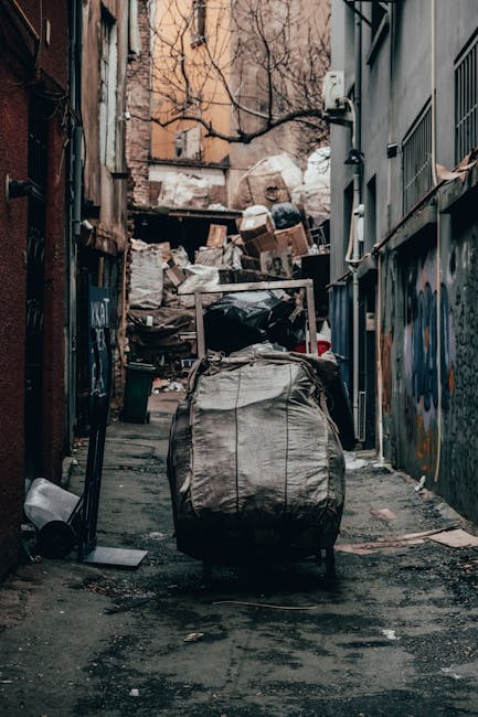 A white commercial van with its rear doors open, positioned on a city street in Fitzrovia, surrounded by large cardboard boxes, plastic-wrapped furniture, and black garbage bags loaded into the vehicle for a house relocation or bulky waste removal. The boxes are stacked on top of the van, some partially collapsed, with packaging materials visible. A hand truck or dolly is placed beside the open van, loaded with additional waste materials, ready for collection or transport. The background features a multi-storey building with beige, classical-style architecture and several windows. Natural daylight illuminates the scene, highlighting the process of loading waste or unwanted items as part of a professional removal or clearance service provided by Man with Van Fitzrovia, supporting efficient moving logistics and packing and moving operations.