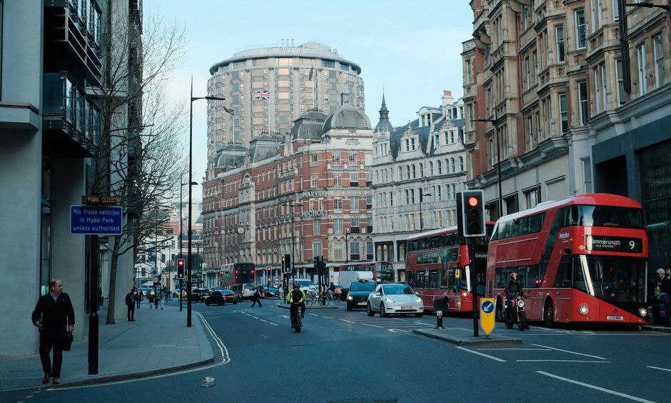 A narrow urban street scene in Fitzrovia, showing a row of historic brick buildings with arched windows and decorative architectural details on the left side, and a modern glass-fronted building refle