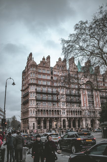 A multi-storey historic building with elaborate stone architecture and large windows, situated on a busy urban street with parked cars and moving vehicles. In the foreground, several pedestrians are w