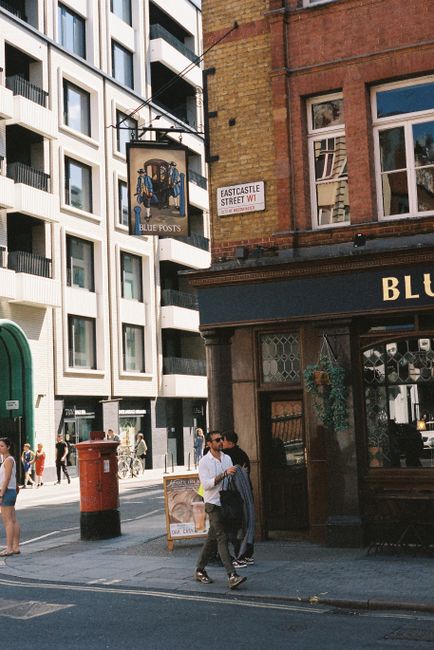 Image of a busy street scene in Fitzrovia with a move-related context, showing front views of a modern multi-story building with numerous windows on the left and a traditional brick building with large windows and a storefront named 'Blue Post' on the right. The storefront has a dark awning, potted plants, and decorative window features, indicating a commercial premise. In the foreground, two men are walking on the pavement, with one carrying a backpack and the other holding a paper shopping bag, while other pedestrians are visible in the background. A red post box is positioned near the curb, and an street sign reading 'Eastcastle Street W1' is mounted on the brick building. The scene captures everyday urban activity consistent with home or business relocations, possibly involving packing, furniture transport, or moving logistics with a focus on Fitzrovia. The lighting suggests daytime, with natural sunlight illuminating the scene, and the setting appears suitable for a UK-based removals or moving service like Man with Van Fitzrovia.