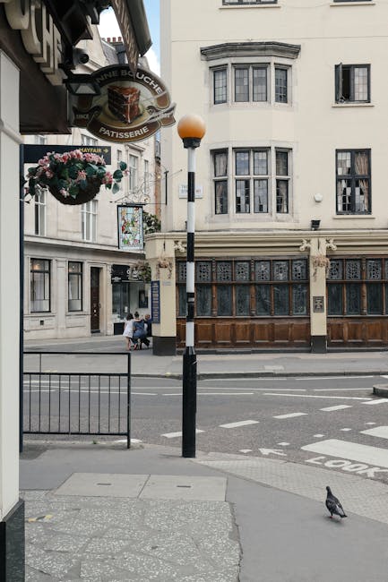 Image of a busy street scene in Fitzrovia with a move-related context, showing front views of a modern multi-story building with numerous windows on the left and a traditional brick building with large windows and a storefront named 'Blue Post' on the right. The storefront has a dark awning, potted plants, and decorative window features, indicating a commercial premise. In the foreground, two men are walking on the pavement, with one carrying a backpack and the other holding a paper shopping bag, while other pedestrians are visible in the background. A red post box is positioned near the curb, and an street sign reading 'Eastcastle Street W1' is mounted on the brick building. The scene captures everyday urban activity consistent with home or business relocations, possibly involving packing, furniture transport, or moving logistics with a focus on Fitzrovia. The lighting suggests daytime, with natural sunlight illuminating the scene, and the setting appears suitable for a UK-based removals or moving service like Man with Van Fitzrovia.