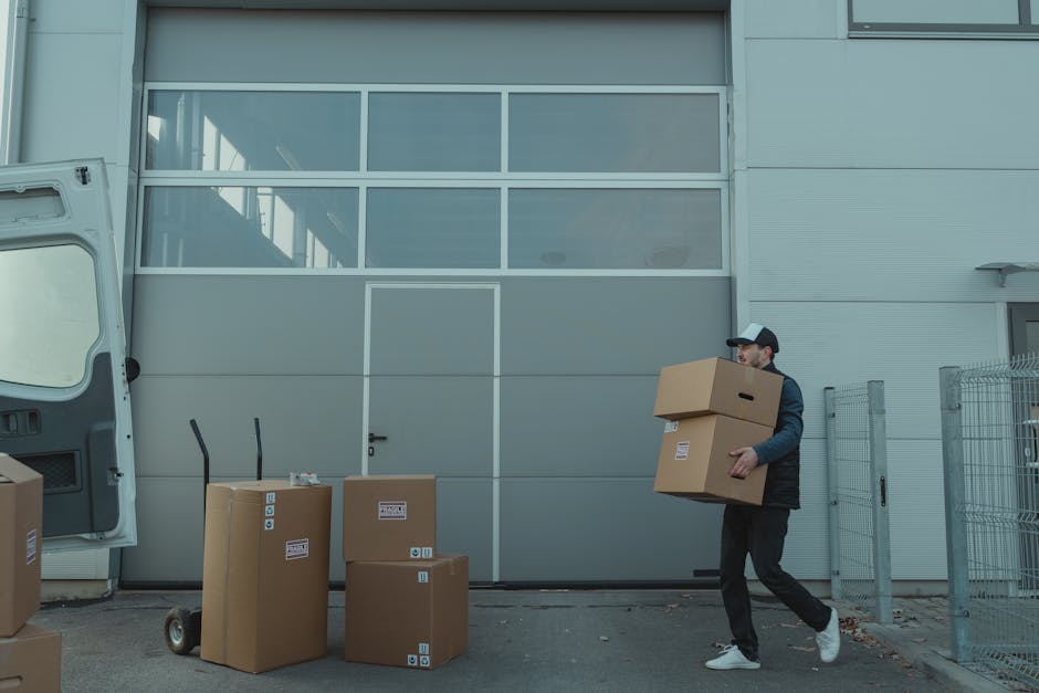 A man wearing casual clothing, including a baseball cap, black jacket, and dark trousers, is carrying two cardboard boxes, one stacked on top of the other, outside a modern, grey industrial building w