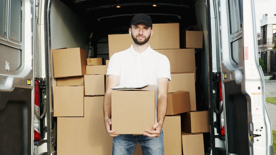A man wearing casual clothing, including a baseball cap, black jacket, and dark trousers, is carrying two cardboard boxes, one stacked on top of the other, outside a modern, grey industrial building w