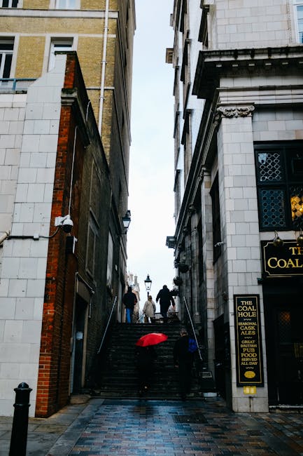 A narrow outdoor street in Fitzrovia viewed from street level, showing a steep set of stone stairs with a metal handrail on each side, leading upward between two multi-storey buildings. On the stairs, several people are seen carrying or walking past large items covered with blankets and plastic wrapping, possibly during a home relocation or moving process. Some smaller cardboard boxes and packing materials are visible on the steps. To the left, the building features a mix of brick and white paneled facades, while to the right, the building has a white exterior with ornate architectural details and a sign for 'The Coal Hole' pub. An umbrella is placed on the ground near the base of the stairs, and the surrounding environment appears wet, reflecting recent rain, with overcast skies above. The scene illustrates a typical move involving furniture transport through a narrow, urban Fitzrovia street, supported by [COMPANY_NAME]'s removals services.