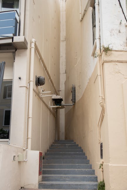 A narrow outdoor staircase with dark grey steps ascending between two tall, beige building walls. The walls feature visible piping, electrical conduit, and a small black box mounted on the left side, along with some minor peeling paint and small patches of greenery growing from crevices. The setting appears to be an alleyway used for access to upper floors or for moving purposes. This image relates to home relocation and furniture transport, highlighting the challenges of navigating narrow spaces during packing and moving activities, as seen in services provided by Man with Van Fitzrovia.