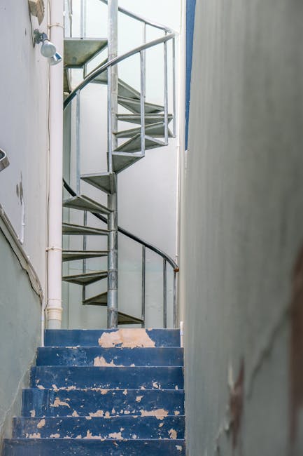 A narrow outdoor street in Fitzrovia viewed from street level, showing a steep set of stone stairs with a metal handrail on each side, leading upward between two multi-storey buildings. On the stairs, several people are seen carrying or walking past large items covered with blankets and plastic wrapping, possibly during a home relocation or moving process. Some smaller cardboard boxes and packing materials are visible on the steps. To the left, the building features a mix of brick and white paneled facades, while to the right, the building has a white exterior with ornate architectural details and a sign for 'The Coal Hole' pub. An umbrella is placed on the ground near the base of the stairs, and the surrounding environment appears wet, reflecting recent rain, with overcast skies above. The scene illustrates a typical move involving furniture transport through a narrow, urban Fitzrovia street, supported by [COMPANY_NAME]'s removals services.