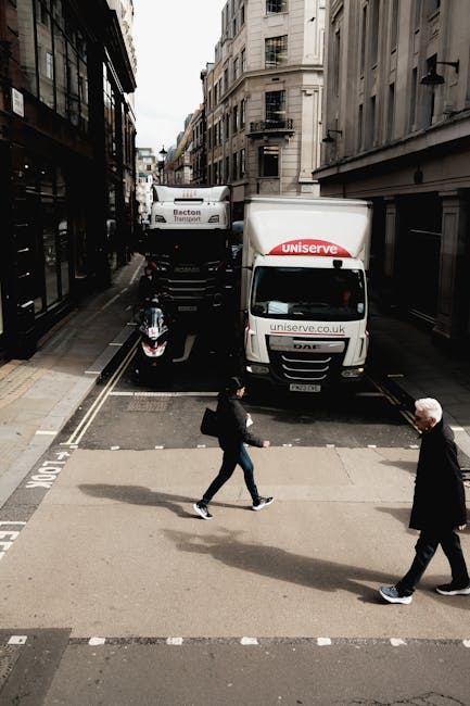 A street scene showing a narrow urban road lined with tall, multi-storey buildings with traditional facades. Two large white moving trucks with branding from Uniserve and Becton Transport are parked along the curb, partially blocking the road. One of the trucks is positioned at an angle, suggesting recent or ongoing loading or unloading activities related to home relocation or furniture transport. A motorcycle is parked between the trucks on the road. In the foreground, two pedestrians are crossing the street at a pedestrian crossing; one is a woman carrying a bag, and the other is an older man with white hair, both dressed in dark clothing. Soft natural light illuminates the scene, capturing the busy environment typical of a city centre, with the distance showing additional buildings and a narrow alleyway. This image reflects general moving logistics and professional removals activities.
