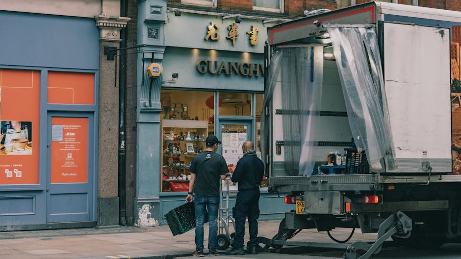 Two movers from Man with Van Fitzrovia are preparing to load a small moving truck parked outside a shop on a city street. The truck has its side panel lifted, revealing a partially open plastic curtain that conceals the interior for furniture transport and packing. One mover, dressed in dark clothing, is standing next to a hand truck, while the other, wearing a black shirt and jeans, is holding a plastic crate. They are positioned on the pavement at the entrance of a shop named Guangh, which has a grey and orange exterior with Chinese characters and the word 'GUANGH' visible above the doorway. Inside the shop, various items such as decorative objects and appliances are visible through the glass window, indicating a retail establishment. The scene captures a typical home relocation or furniture transport process, with the movers preparing goods for loading during a logistics operation associated with house removals and packing services. Natural daylight illuminates the scene, emphasizing the urban environment and the professional activity of loading items for a house move.