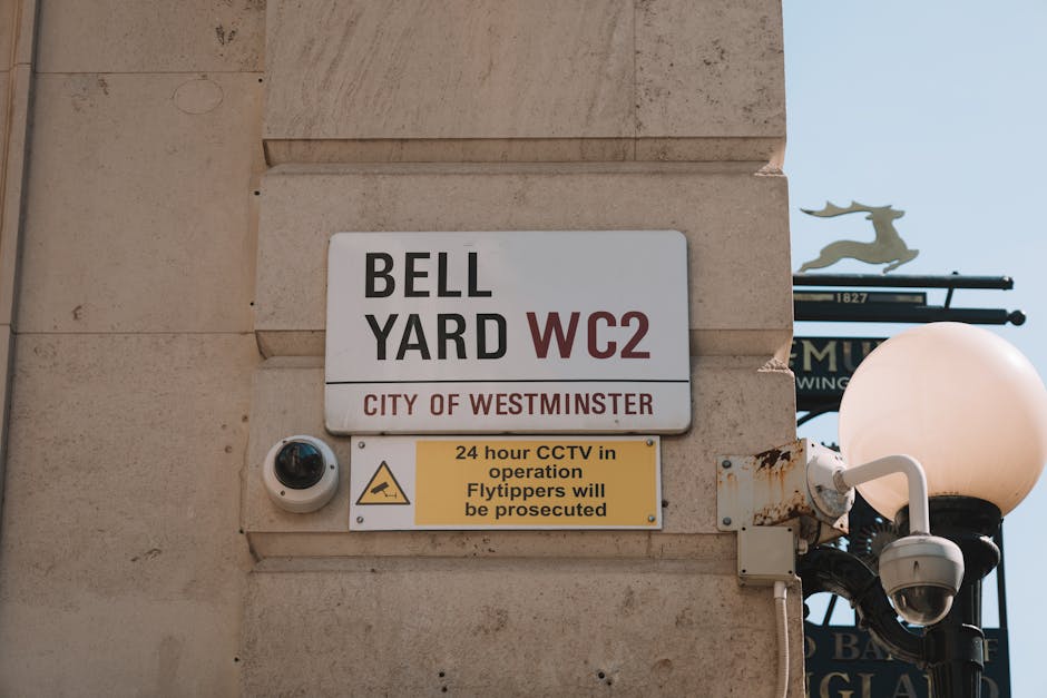 Close-up image of a building corner showing a white and red sign indicating the Bell Yard WC2 parking suspensions for Westminster, UK, with text 'CITY OF WESTMINSTER'. Below the sign, a yellow notice warns that 24-hour CCTV is in operation and fly-tippers will be prosecuted. To the left, a dome-shaped CCTV camera is mounted on the wall, aimed at the street to monitor parking and loading activities. On the right side, part of a decorative street lamp with a white spherical light is visible, attached to a black metal post. The background features a clear blue sky with a tree branch partially visible at the top. The setting suggests the signage is part of the infrastructure supporting house and furniture removals, ensuring designated parking and security during home relocation or moving services carried out by [COMPANY_NAME].