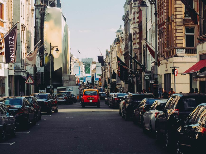 A busy city street scene in Fitzrovia during daylight, with parked cars lining both sides of the road and a narrow lane in the centre. The street is bordered by multi-storey buildings featuring detailed facades, some with flags and signage, and shopfronts on the ground level. On the left, there are several dark-colored vehicles, including sedans and small vans, parked parallel to the pavement. In the middle of the street, a red classic London taxi is seen moving, surrounded by more parked cars. The right side of the image shows additional parked vehicles, including a silver hatchback and a dark SUV. Pedestrians are visible walking along the pavements, and in the background, the street extends into a bustling city area with billboards, streetlights, and signage. The overall scene captures typical urban furniture transport and home relocation activity, consistent with London’s Fitzrovia district, fitting for a housing move or furniture transport managed by Man with Van Fitzrovia during a scheduled parking suspension.